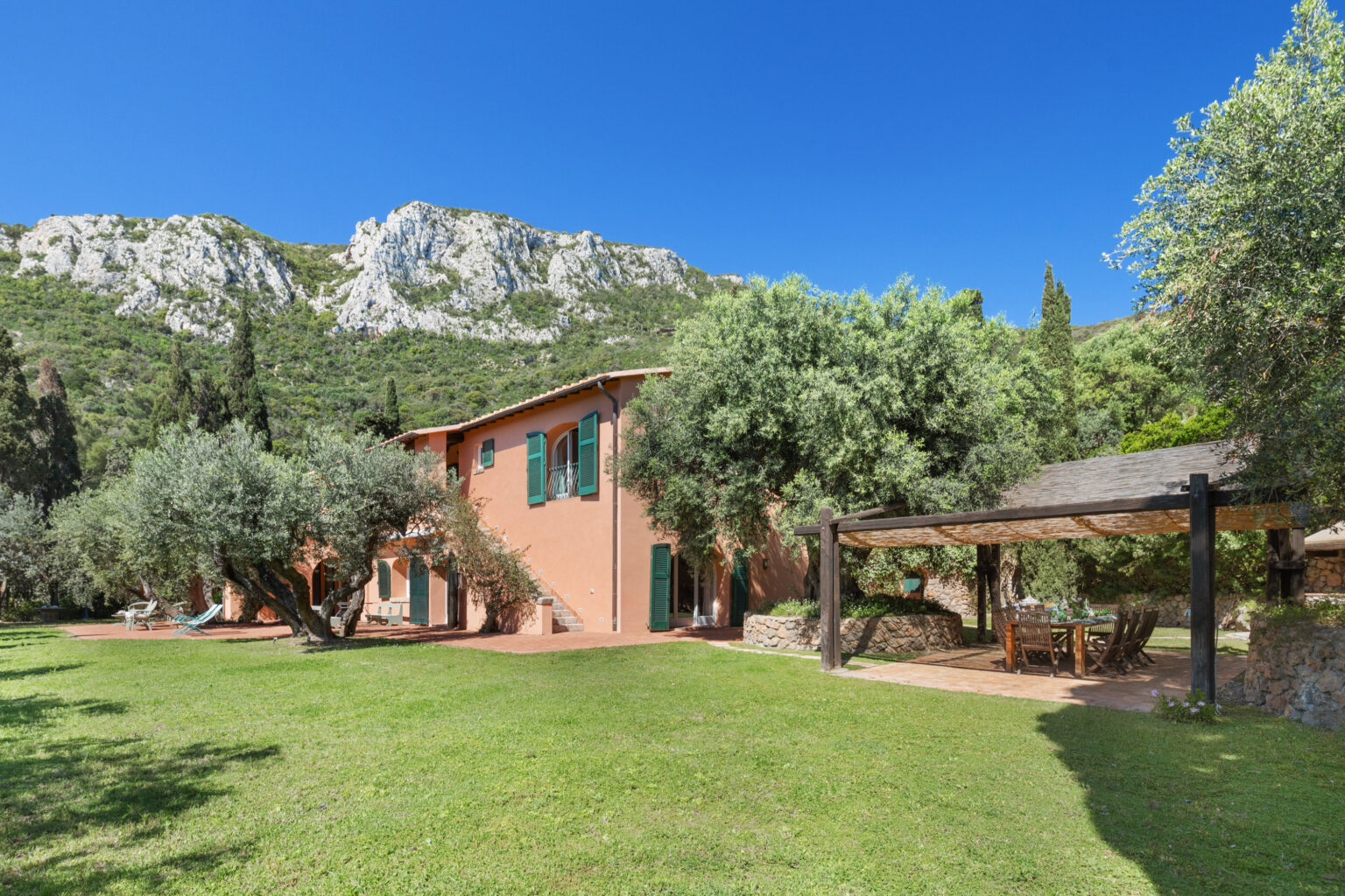 Charming rustic villa surrounded by lush greenery and mountains, featuring a spacious garden, outdoor seating area, and olive trees under a clear blue sky. Ideal for relaxation and nature retreats. | Only on SopranoVillas