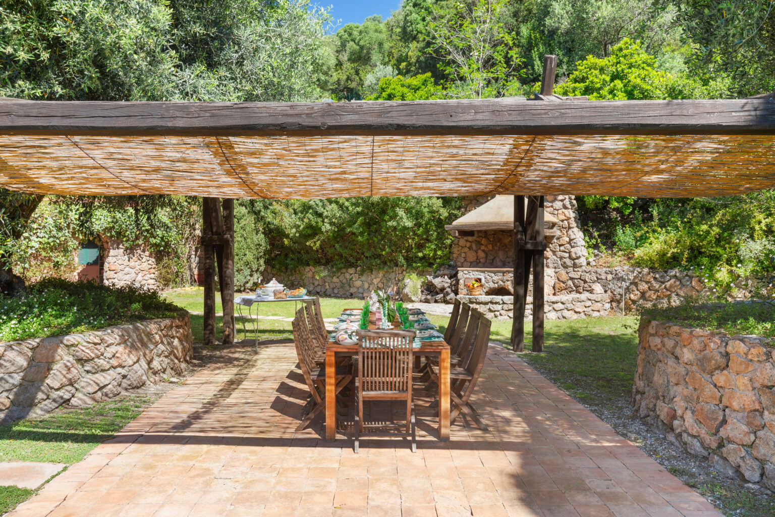 Outdoor dining area with a wooden table set for a meal, surrounded by lush greenery and stone walls, featuring a thatched roof for shade. Ideal for alfresco dining and gatherings in a serene garden setting. | Only on SopranoVillas