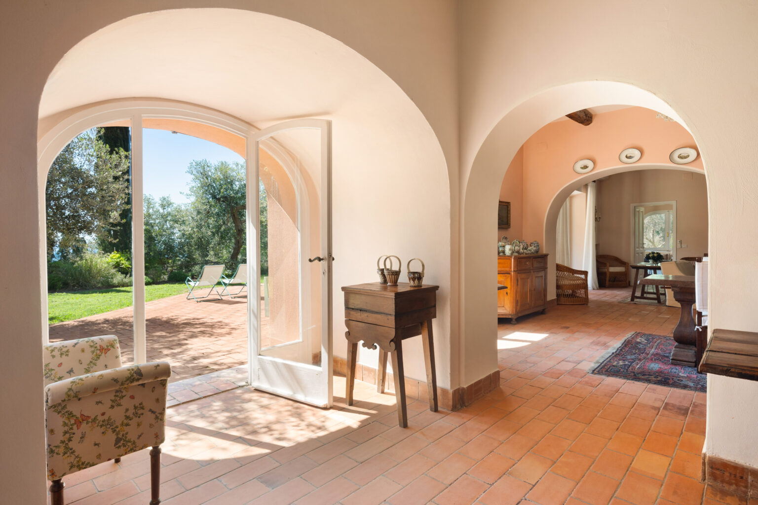 Interior view of a stylish home showcasing arched doorways, terracotta flooring, and a cozy seating area. Natural light floods the space, highlighting the wooden furniture and inviting ambiance, with a glimpse of the lush garden visible through the open doors. | Only on SopranoVillas