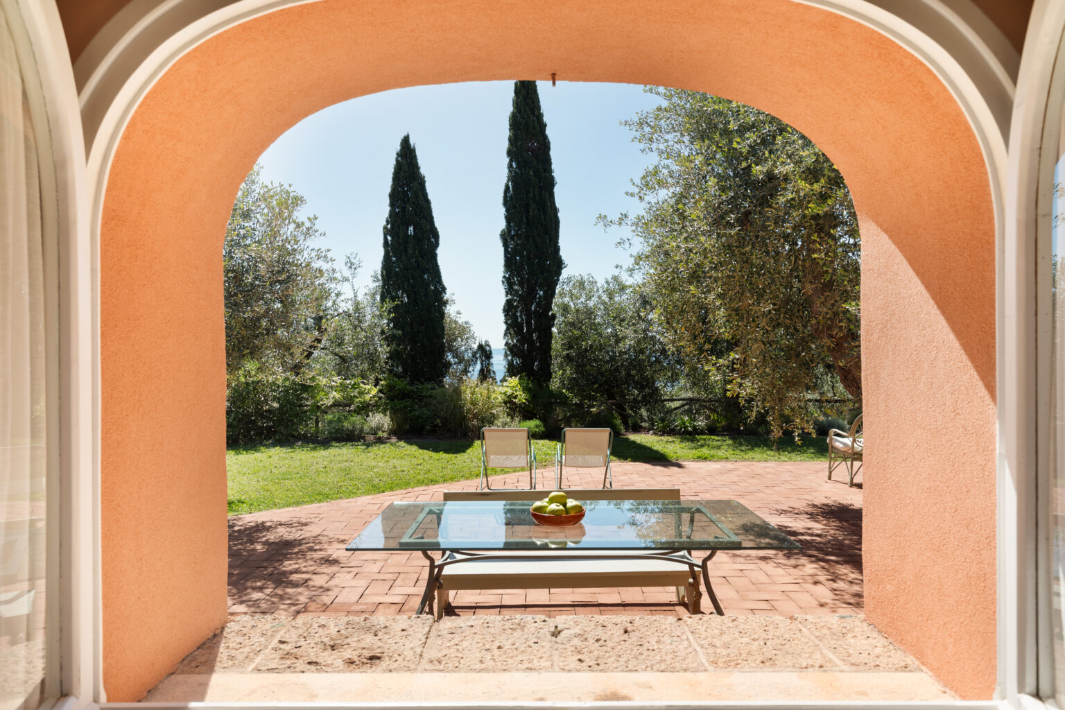 View from an arched window showcasing a serene outdoor space with a glass table and two chairs on a brick patio, surrounded by lush greenery and tall cypress trees under a clear blue sky. | Only on SopranoVillas