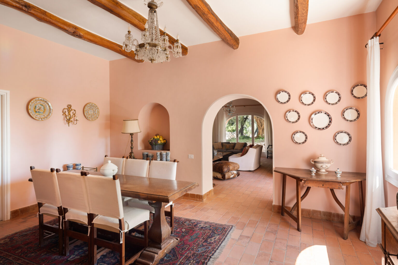 Spacious dining area with a wooden table and upholstered chairs, accented by a chandelier and decorative wall plates. Soft peach walls and terracotta flooring create a warm atmosphere, while an arched doorway leads to a cozy living space. Natural light floods the room through large windows. | Only on SopranoVillas