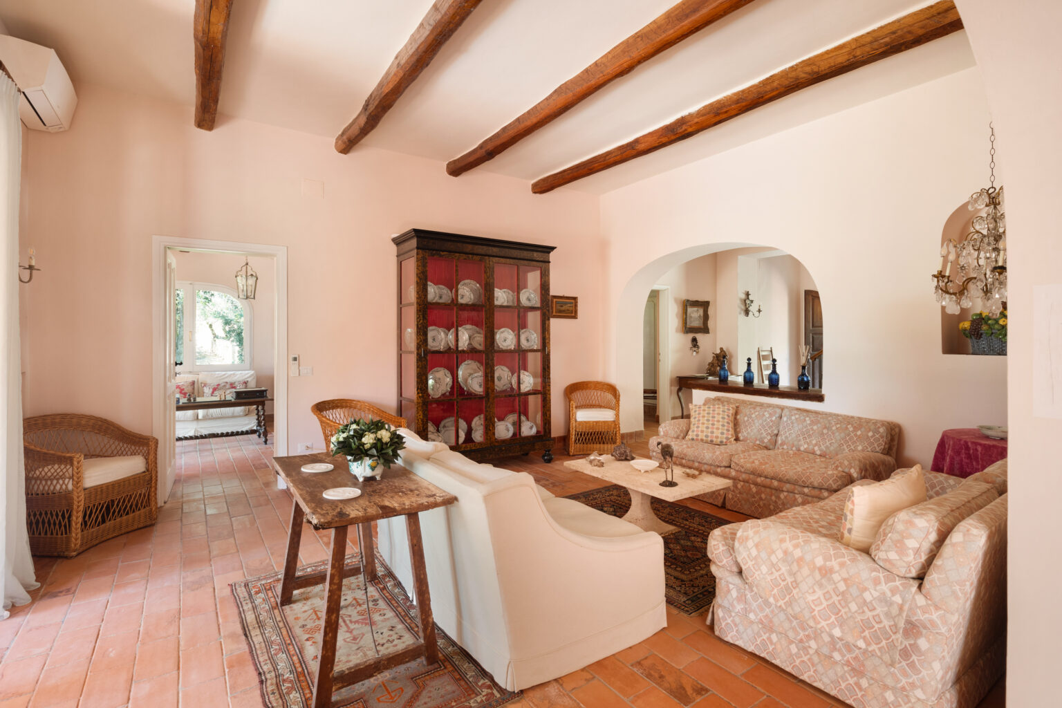Cozy living room featuring rustic wooden beams, a warm color palette, and elegant furnishings. The space includes a vintage wooden table, comfortable couches, and a glass-fronted cabinet displaying decorative plates, creating a welcoming atmosphere. | Only on SopranoVillas
