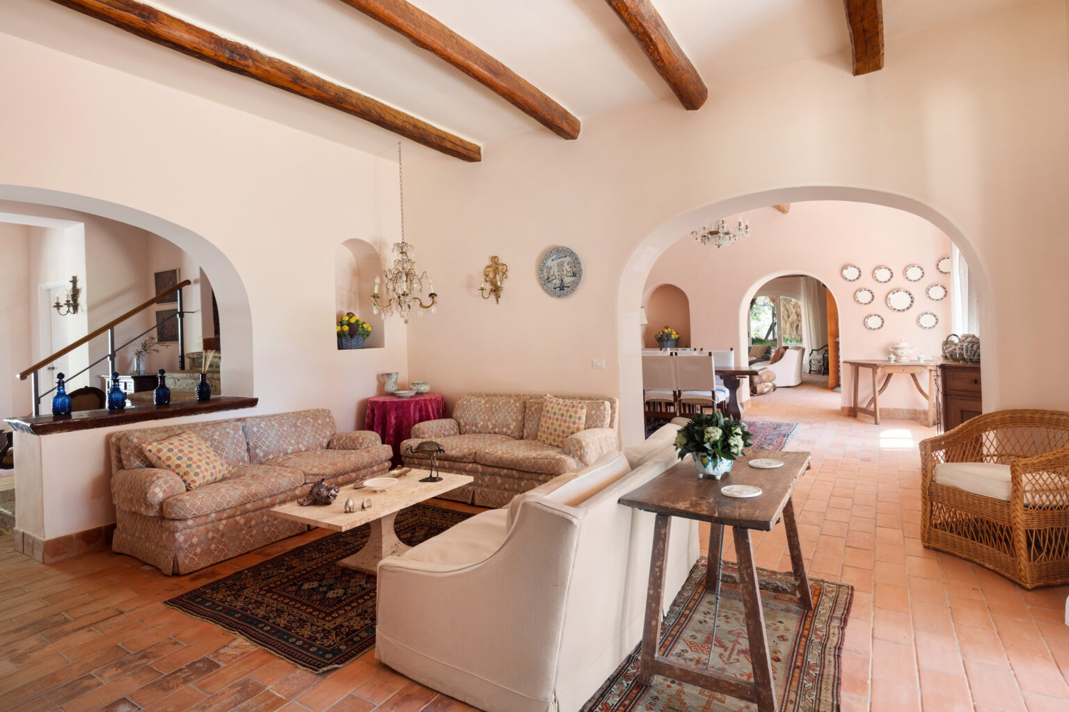 Bright and inviting living room featuring exposed wooden beams, cozy sofas, and a marble coffee table. The space is adorned with decorative elements such as chandeliers and artwork, complemented by terracotta flooring and a warm color palette. Arched doorways lead to a dining area, enhancing the open layout. | Only on SopranoVillas