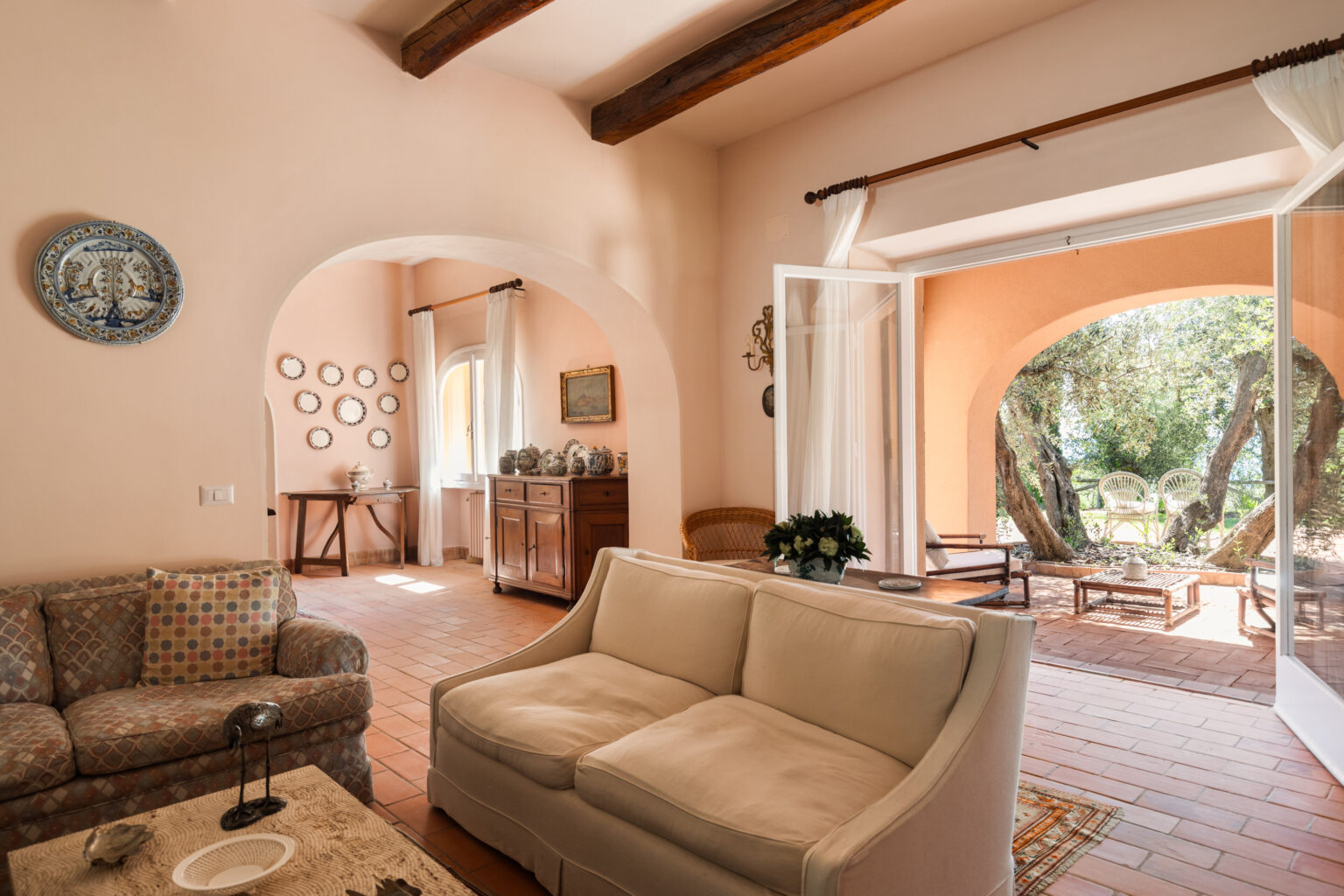 Bright and airy living room featuring a comfortable cream-colored sofa, decorative wall plates, and rustic wooden beams. The room opens to a serene outdoor patio with olive trees and seating, creating a cozy indoor-outdoor living space. | Only on SopranoVillas