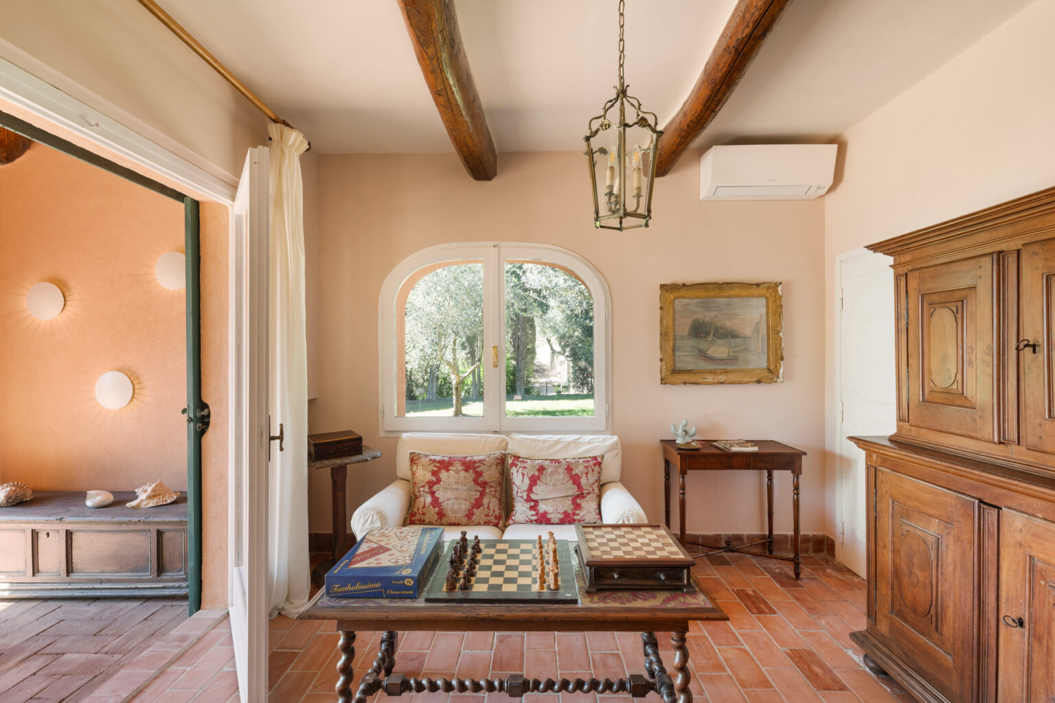 Cozy living room featuring a white sofa with decorative pillows, a wooden table with chess and board games, and rustic wooden furniture. Large windows provide a view of the garden, complemented by warm beige walls and exposed wooden beams. Natural light enhances the inviting atmosphere. | Only on SopranoVillas