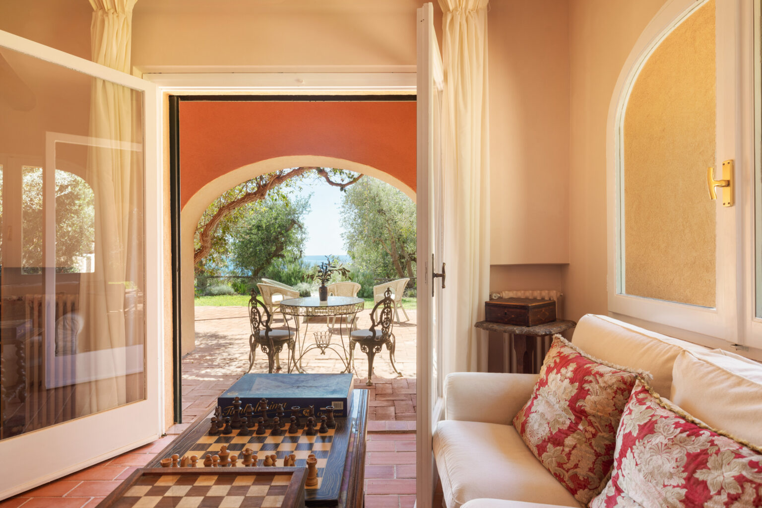 Cozy interior view of a living room with a chessboard, leading to an outdoor patio with a table and chairs surrounded by greenery and a sea view. The space features warm colors, natural light, and elegant furnishings, creating a serene atmosphere. | Only on SopranoVillas