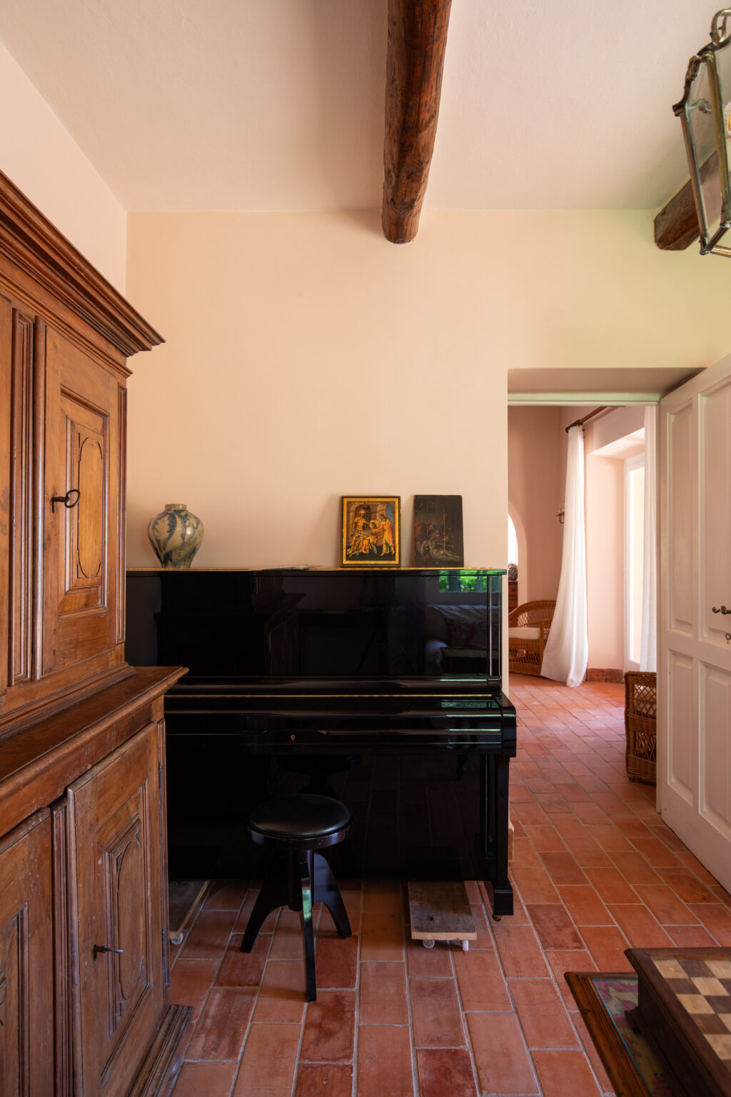 Interior view of a cozy room featuring a black piano with a wooden stool, an antique wooden cabinet, and decorative items including a vase and framed artwork. The terracotta tiled floor and soft lighting create a warm ambiance, with a doorway leading to another room in the background. | Only on SopranoVillas