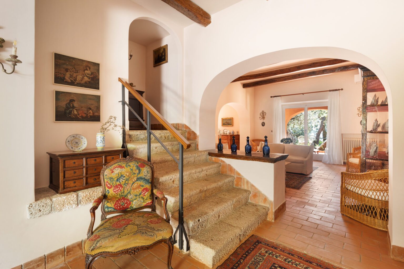 Welcoming entrance hallway featuring a decorative chair, wooden staircase, and vintage artwork on the walls, leading to a cozy living area with natural light and stylish furnishings. | Only on SopranoVillas