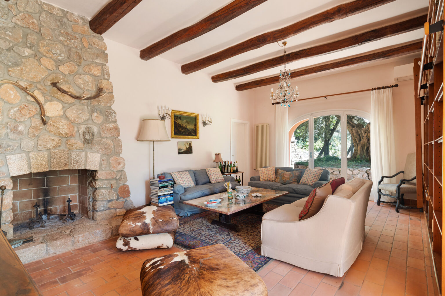Cozy living room featuring a stone fireplace, wooden beams, and comfortable seating. Decor includes cowhide ottomans, a large coffee table, and a chandelier, with natural light flooding in through large windows overlooking a garden. | Only on SopranoVillas