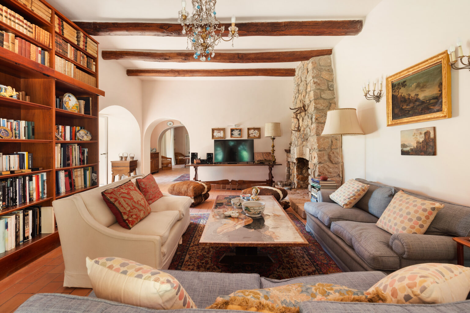Cozy living room featuring a stone fireplace, wooden beams, and a large bookshelf filled with books. The space includes comfortable sofas with decorative cushions, a coffee table with tableware, and tasteful artwork on the walls, creating an inviting atmosphere. | Only on SopranoVillas