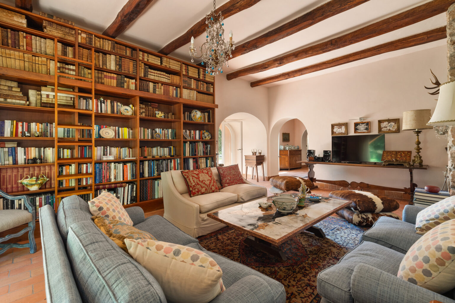 Cozy living room featuring a large wooden bookshelf filled with a variety of books, a plush seating area with decorative pillows, a marble coffee table, and a chandelier. The room has exposed wooden beams and a warm color palette, creating an inviting atmosphere. | Only on SopranoVillas