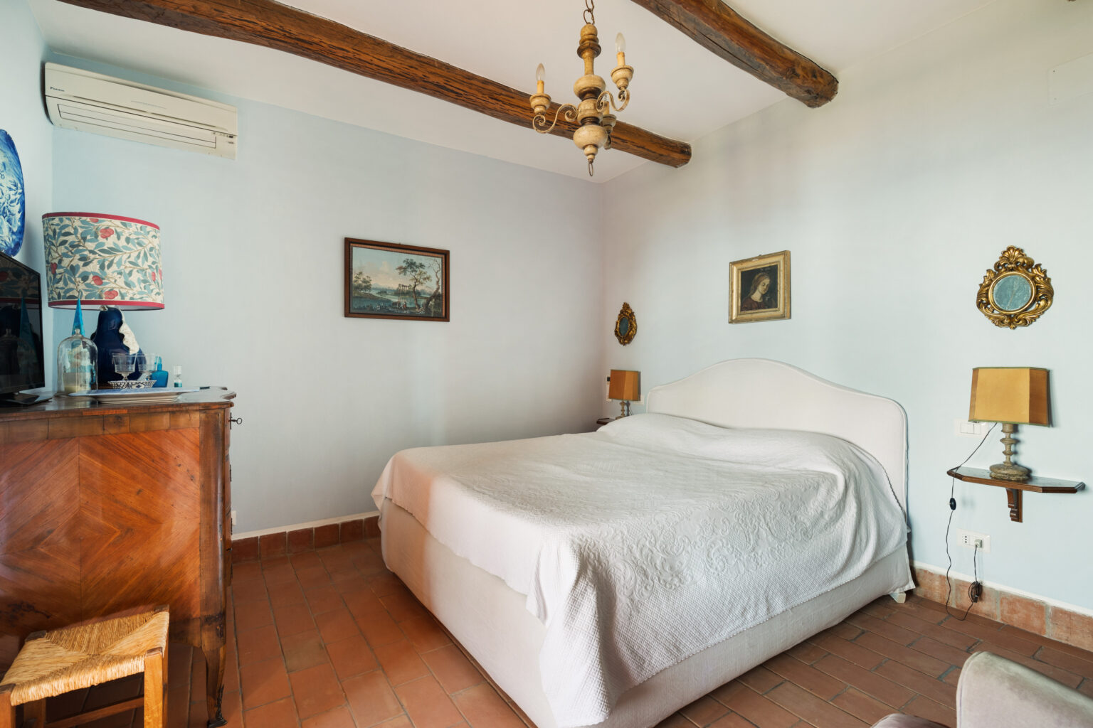 Cozy bedroom featuring a white bedspread, antique wooden furniture, and decorative wall art. The room has exposed wooden beams, a vintage chandelier, and a colorful lamp, creating a warm and inviting atmosphere. | Only on SopranoVillas