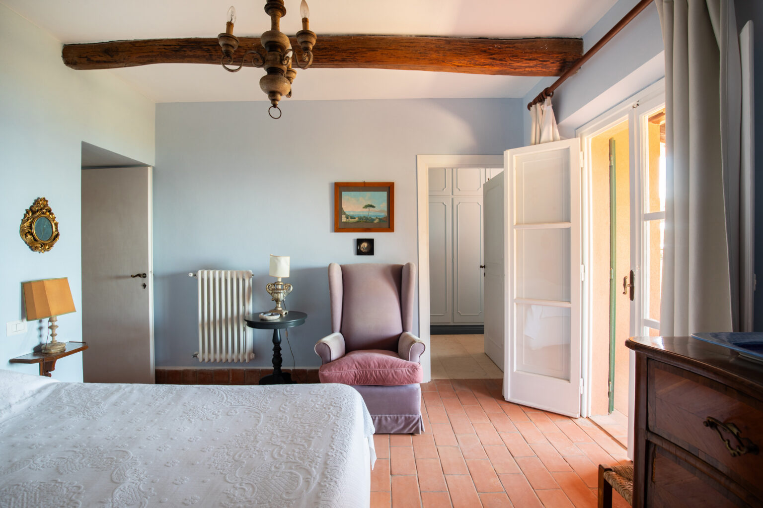 Cozy bedroom interior featuring a comfortable bed with a decorative white bedspread, a vintage chandelier with wooden beams overhead, a plush pink armchair, and a small black side table with a lamp. The room has light blue walls, a window with curtains, and an open door leading to another space, creating a warm and inviting atmosphere. | Only on SopranoVillas