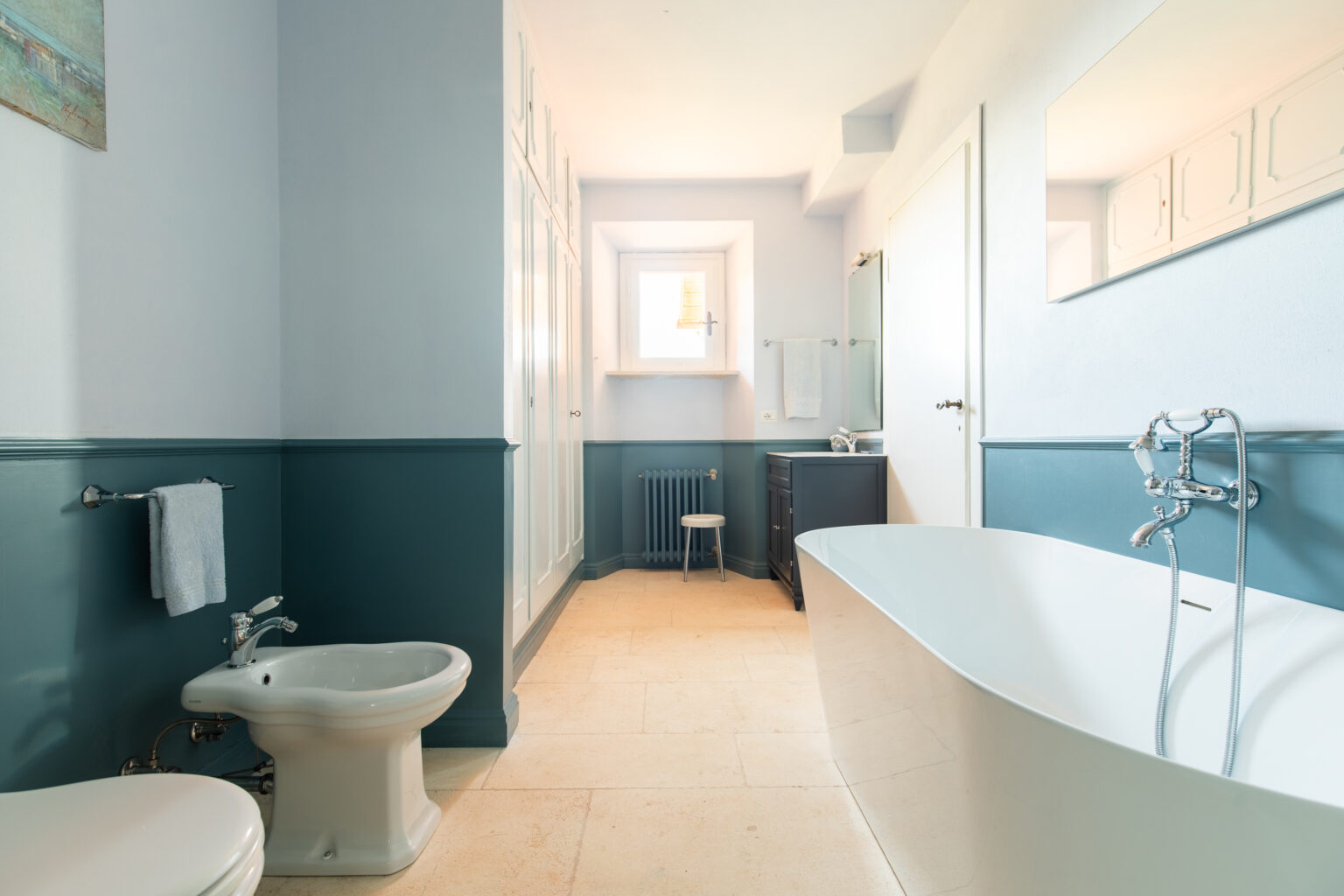 Modern bathroom featuring a freestanding white bathtub, bidet, and stylish cabinetry. The walls are painted in soft blue with a darker blue wainscoting, complemented by natural light from a window. Elegant fixtures and a minimalistic design create a serene atmosphere. | Only on SopranoVillas