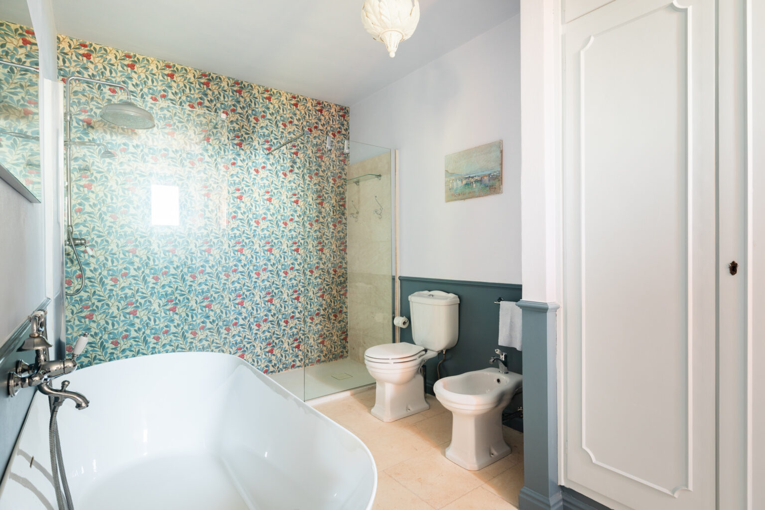 Bright and modern bathroom featuring a freestanding white bathtub, a walk-in shower with glass doors, and floral wallpaper. The space includes a toilet and bidet, complemented by soft lighting and a decorative painting on the wall. | Only on SopranoVillas