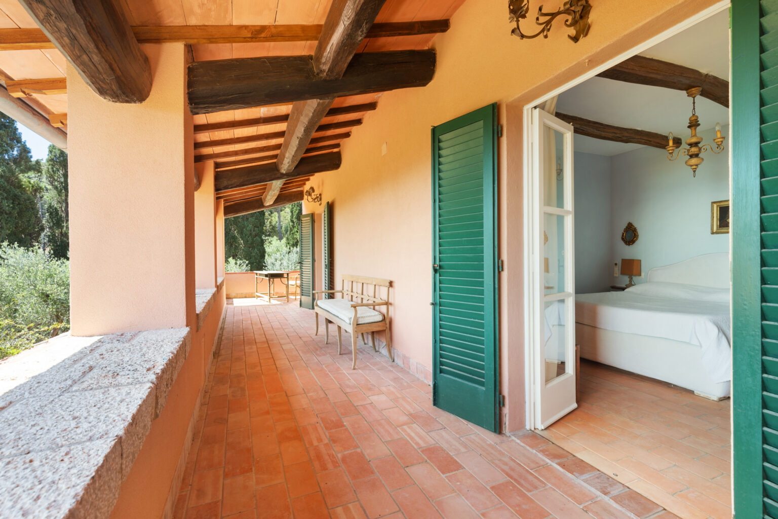 Spacious terrace of a charming villa featuring rustic wooden beams, terracotta flooring, and green shutters. The view leads to a cozy bedroom with a white bed and decorative accents, surrounded by lush greenery. Ideal for relaxation and enjoying nature. | Only on SopranoVillas