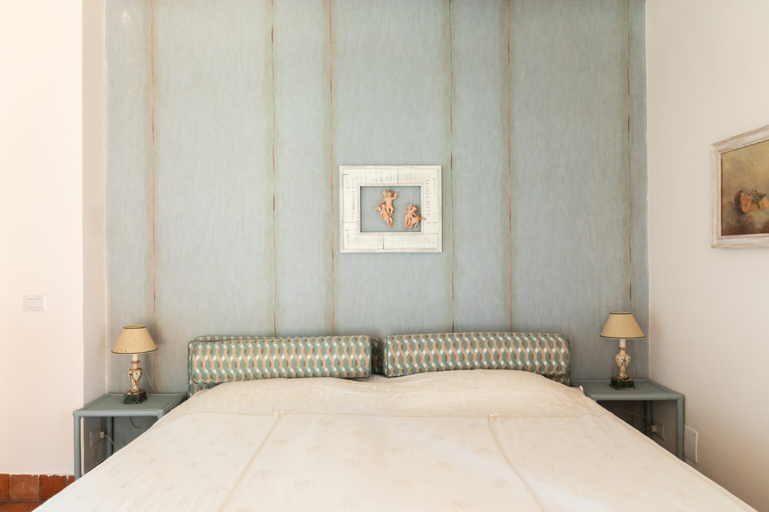 Cozy bedroom featuring a soft, cream-colored bedspread, elegant headboard with a geometric pattern, and decorative wall art. The light blue textured wall creates a serene atmosphere, complemented by bedside lamps and a vintage painting. Perfect for relaxation and comfort in a stylish setting. | Only on SopranoVillas