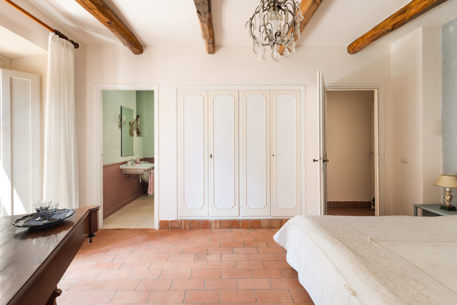 Bright and spacious bedroom featuring terracotta tiled flooring, wooden beam ceiling, and elegant chandelier. The room includes a comfortable bed, a wooden table with decorative plates, and a doorway leading to a modern bathroom with green accents. Ample storage is provided by white built-in wardrobes. | Only on SopranoVillas