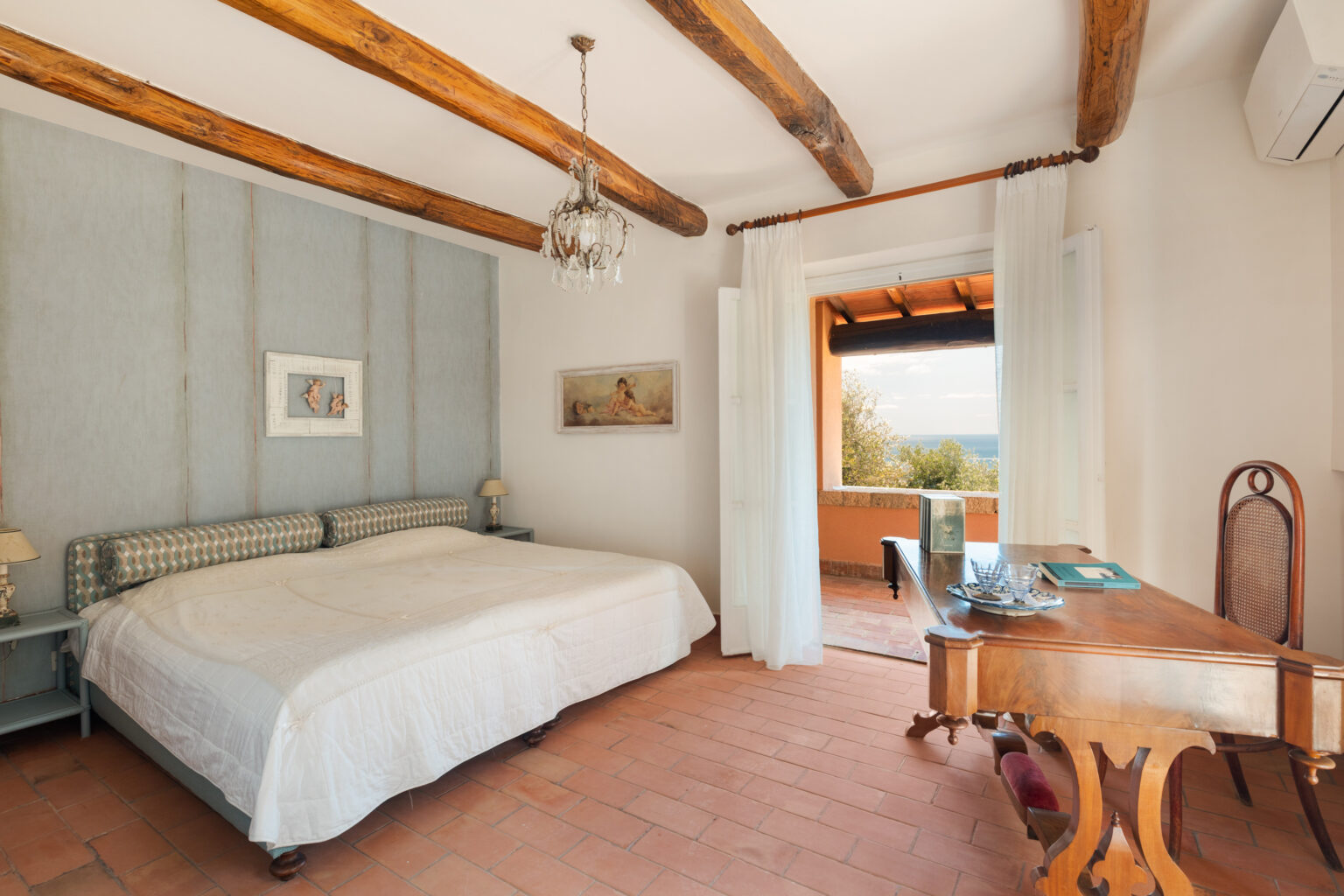 Spacious bedroom featuring wooden beams, a king-sized bed with a patterned headboard, and elegant decor. The room includes a writing desk, soft lighting from bedside lamps, and a large window with a view of the ocean and greenery outside. | Only on SopranoVillas