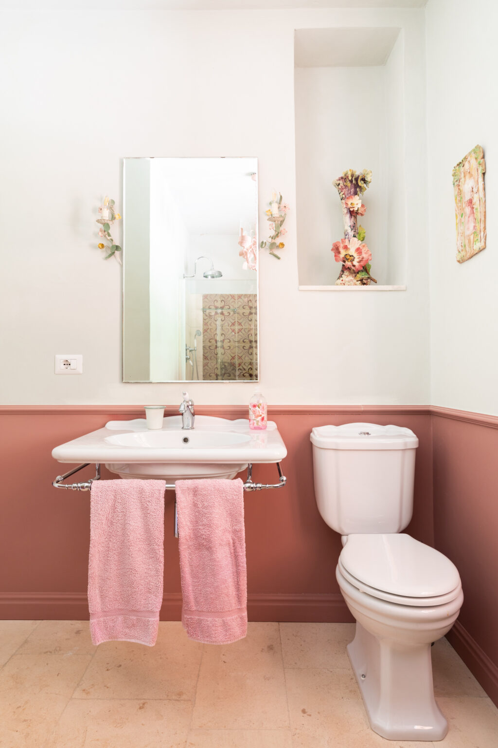 Modern bathroom featuring a white sink with a chrome towel rack holding two pink towels, a white toilet, and a decorative mirror. The walls are painted in soft colors with a floral accent, and a shower area is visible in the background. | Only on SopranoVillas