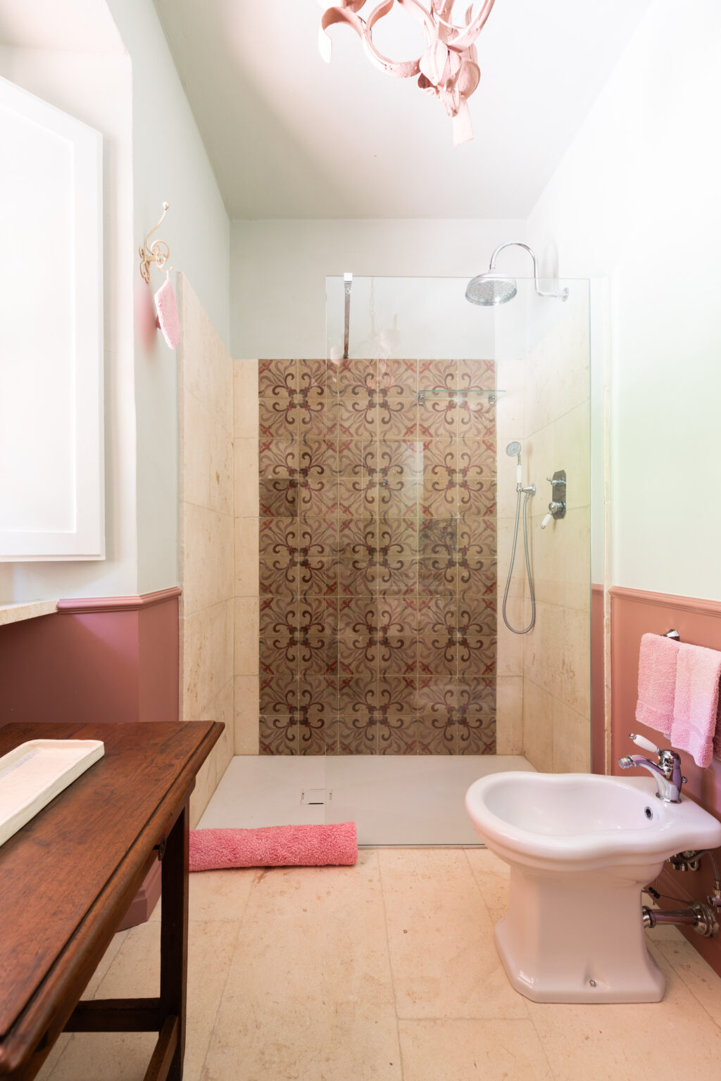 Modern bathroom featuring a glass shower with decorative tile, a wooden vanity, and pink accents. The space includes a bidet, a pink towel, and a stylish chandelier, creating a chic and inviting atmosphere. | Only on SopranoVillas