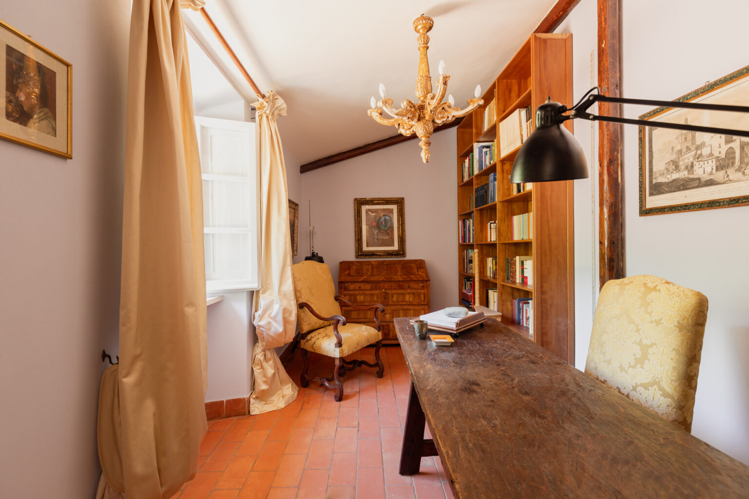 Cozy home office featuring a wooden desk, an ornate chandelier, and a bookshelf filled with books. Natural light streams through the open window, illuminating the elegant decor, including a vintage armchair and framed artwork on the walls. | Only on SopranoVillas
