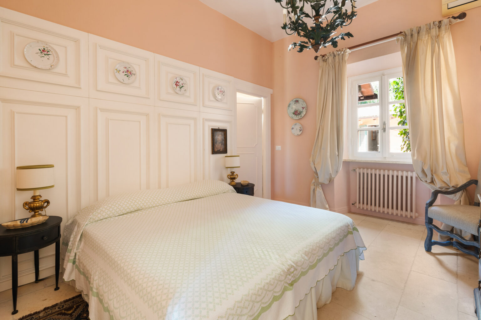 Cozy bedroom featuring a light green bedspread, elegant wall decor with floral plates, and soft peach walls. The room includes a vintage bedside lamp, a small black side table, and large windows with flowing curtains, providing a warm and inviting atmosphere. | Only on SopranoVillas