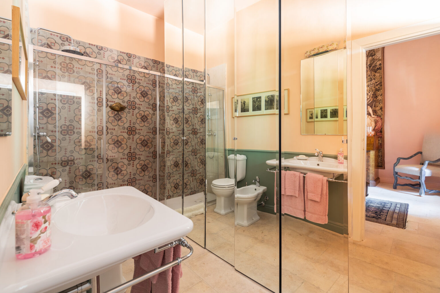 Bright and spacious bathroom featuring a glass shower enclosure, modern sink with floral soap, and decorative tile accents. Soft pink towels hang by the sink, and a green and beige color scheme adds a soothing ambiance. Natural light enhances the room's inviting atmosphere. | Only on SopranoVillas