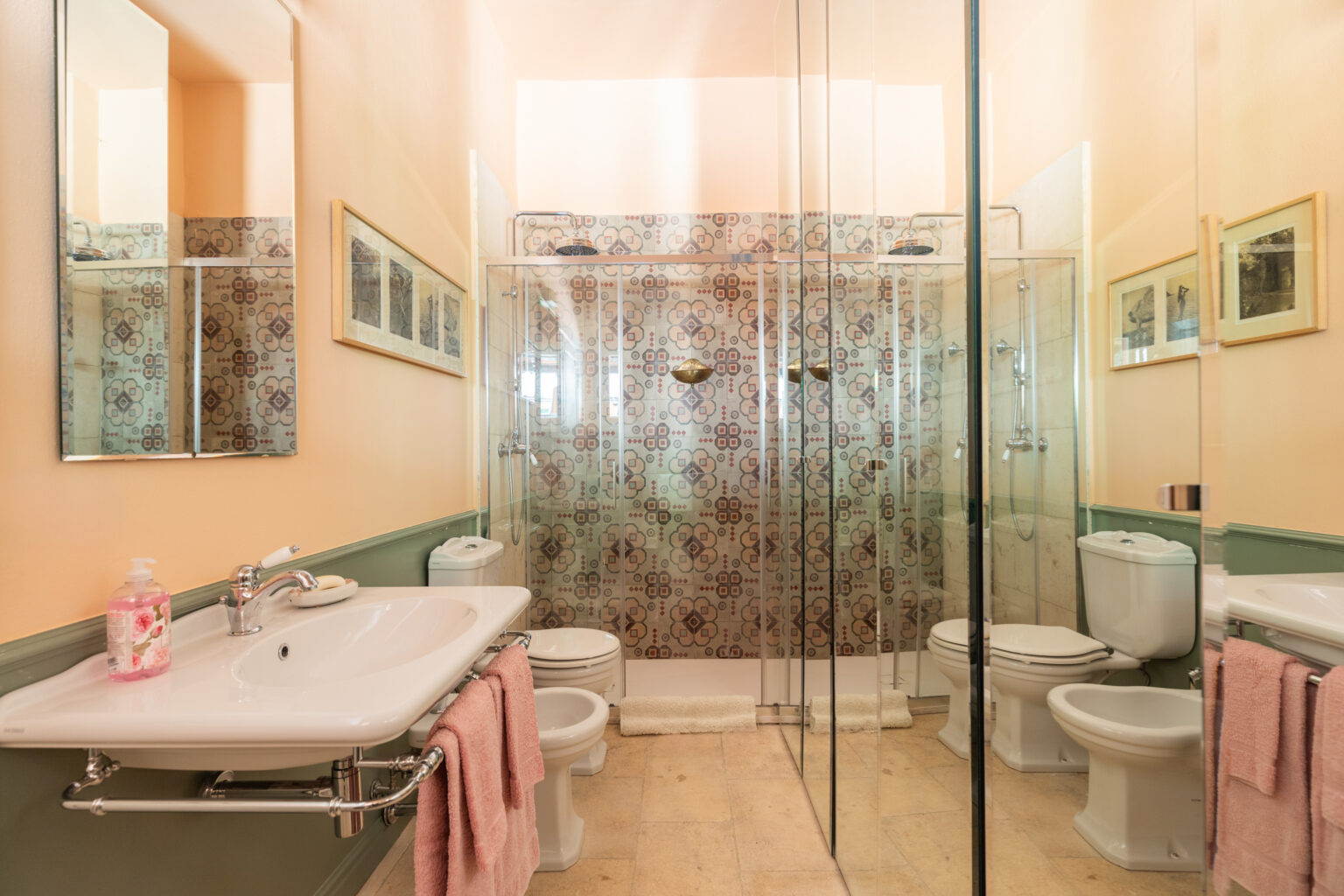 Modern bathroom featuring a glass shower enclosure, stylish patterned tiles, and elegant fixtures. The space includes a white sink with a sleek mirror, a toilet, and a bidet, complemented by soft pink towels and a decorative touch of framed artwork on the walls. | Only on SopranoVillas