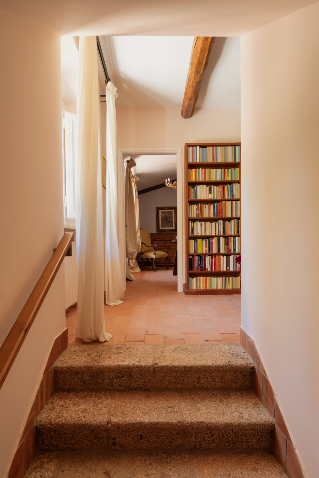 Interior hallway leading to a cozy living area, featuring a bookshelf filled with colorful books, terracotta tiled flooring, and soft curtains hanging from the ceiling. | Only on SopranoVillas