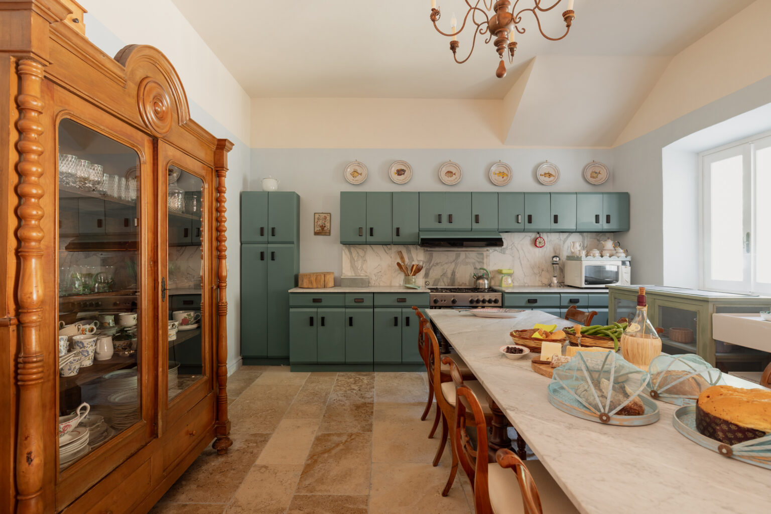 Spacious kitchen featuring green cabinetry, a marble countertop, and a wooden dining table with assorted food items. A vintage wooden cabinet displays fine china, while a chandelier adds a touch of elegance to the room. Natural light streams in through large windows, enhancing the inviting atmosphere. | Only on SopranoVillas
