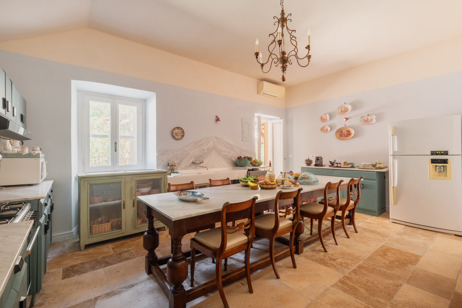Spacious kitchen interior featuring a large wooden dining table surrounded by chairs, a vintage chandelier, and light blue walls. The kitchen includes a modern refrigerator, a stylish cabinet, and decorative plates on the wall, creating a warm and inviting atmosphere. Natural light streams in through the window, enhancing the cozy ambiance. | Only on SopranoVillas