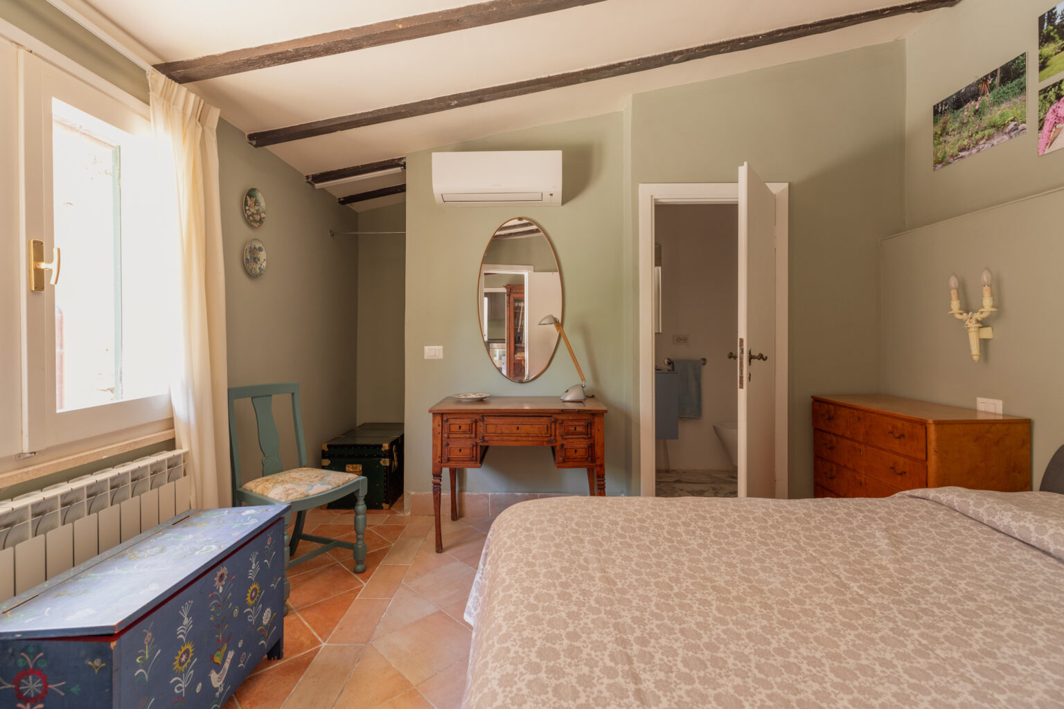 Cozy bedroom featuring a double bed with patterned bedding, rustic wooden beams, and a decorative wall mirror. The room includes a vintage wooden desk, a blue painted storage chest, and a chair with floral upholstery. Natural light streams through a window with sheer curtains, enhancing the inviting atmosphere. A glimpse of an en-suite bathroom is visible through an open door. | Only on SopranoVillas