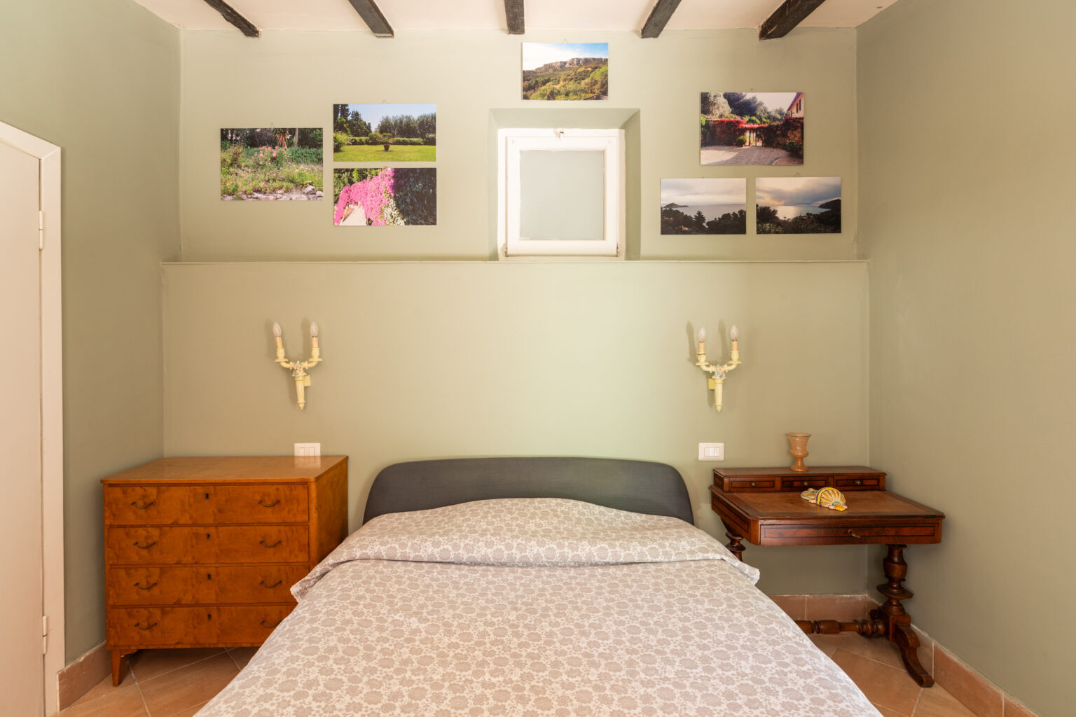 Cozy bedroom featuring a green wall with framed nature photos, a comfortable bed with patterned bedding, and vintage wooden furniture including a nightstand and dresser. Soft lighting from wall sconces enhances the inviting atmosphere. | Only on SopranoVillas