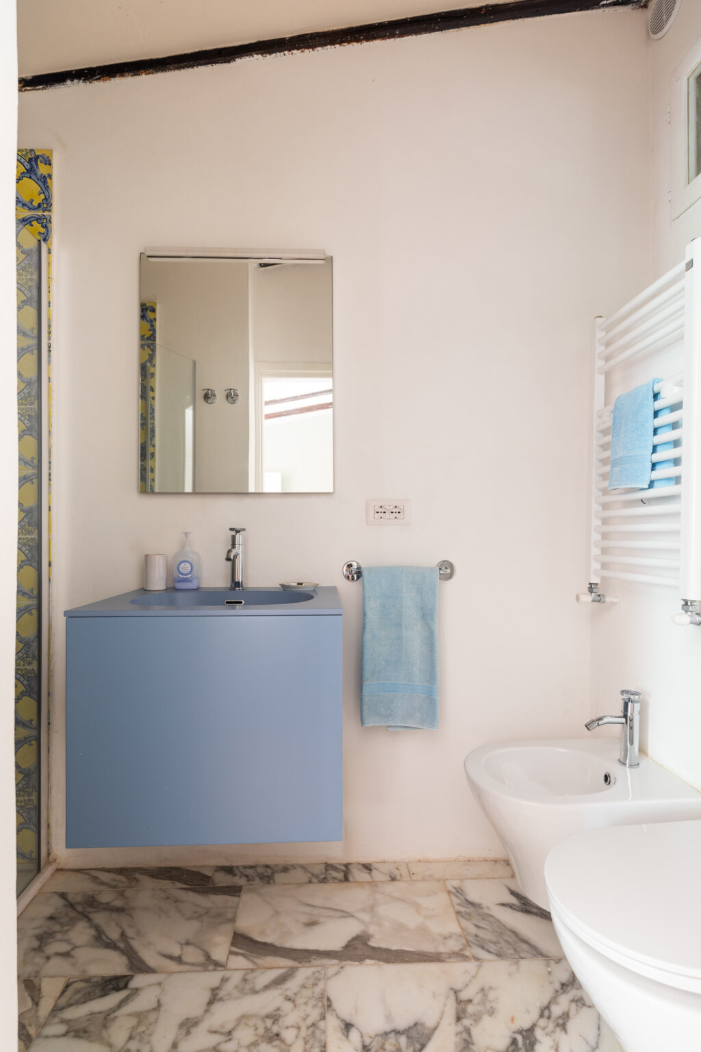 Modern bathroom featuring a blue floating vanity with a sink, a large mirror, and marble flooring. The space includes a bidet, a white toilet, and neatly arranged blue towels on a towel rack, complemented by decorative tiles on the wall. | Only on SopranoVillas