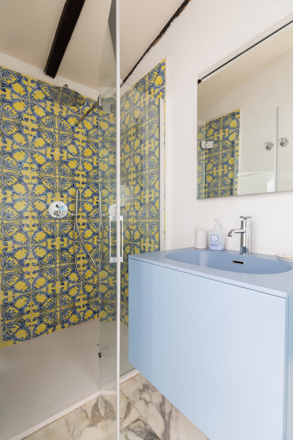 Modern bathroom interior featuring a glass shower enclosure with vibrant blue and yellow patterned tiles, a sleek light blue vanity with a sink, and a marble floor. The design combines functionality with contemporary aesthetics, ideal for a stylish home. | Only on SopranoVillas