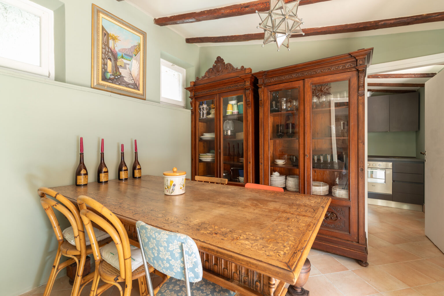 Rustic dining area featuring a vintage wooden table and rattan chairs, with decorative wine bottles and a ceramic jar on the table. Behind the table, there are two elegant glass-front cabinets displaying glassware and tableware, complemented by a colorful landscape painting on the wall. The space has a warm, inviting atmosphere with natural light from the windows and exposed wooden beams on the ceiling. | Only on SopranoVillas