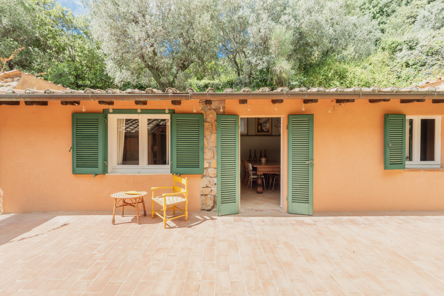 Charming rustic house exterior with green shutters, featuring a cozy porch with a yellow chair and a wicker table, surrounded by lush greenery and a blue sky. Ideal for a peaceful getaway. | Only on SopranoVillas