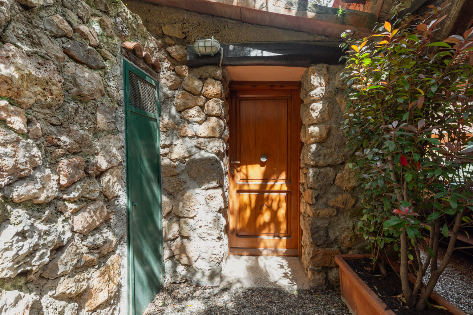 Rustic stone wall featuring a wooden door and a green side door, surrounded by lush greenery and natural light filtering through, creating a warm and inviting entrance. | Only on SopranoVillas