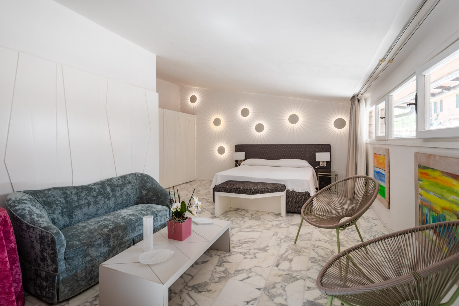 Modern hotel room featuring a stylish design with a plush green sofa, a cozy bed with decorative pillows, and unique wall lighting. The marble floor adds elegance, complemented by colorful artwork and contemporary furniture. Ideal for relaxation and comfort. | Only on SopranoVillas
