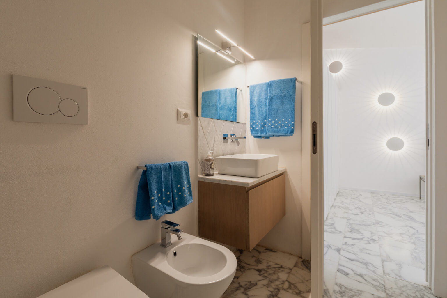 Modern bathroom interior featuring a sleek design with marble flooring, a wall-mounted wooden vanity, a white vessel sink, and stylish blue towels. The space is illuminated by a contemporary light fixture and showcases a bidet. | Only on SopranoVillas