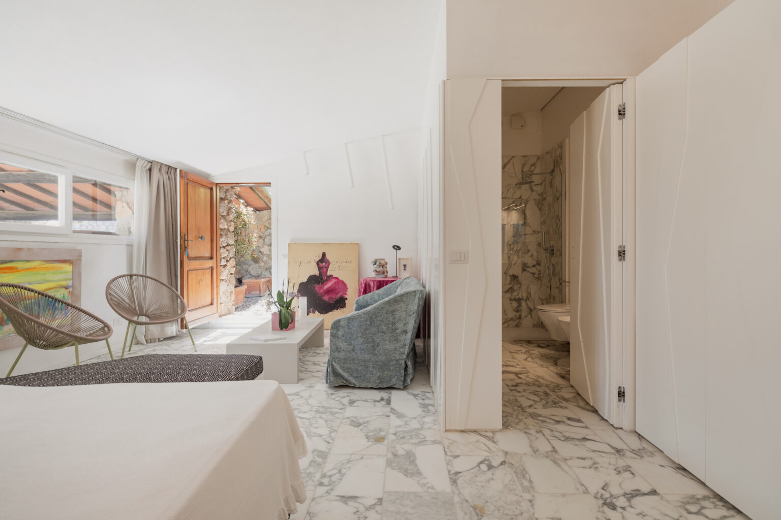 Bright and modern interior of a stylish living space featuring a marble floor, comfortable seating, and an open door leading to an outdoor area. The room includes artistic decor, large windows providing natural light, and a glimpse of a bathroom through a doorway. | Only on SopranoVillas