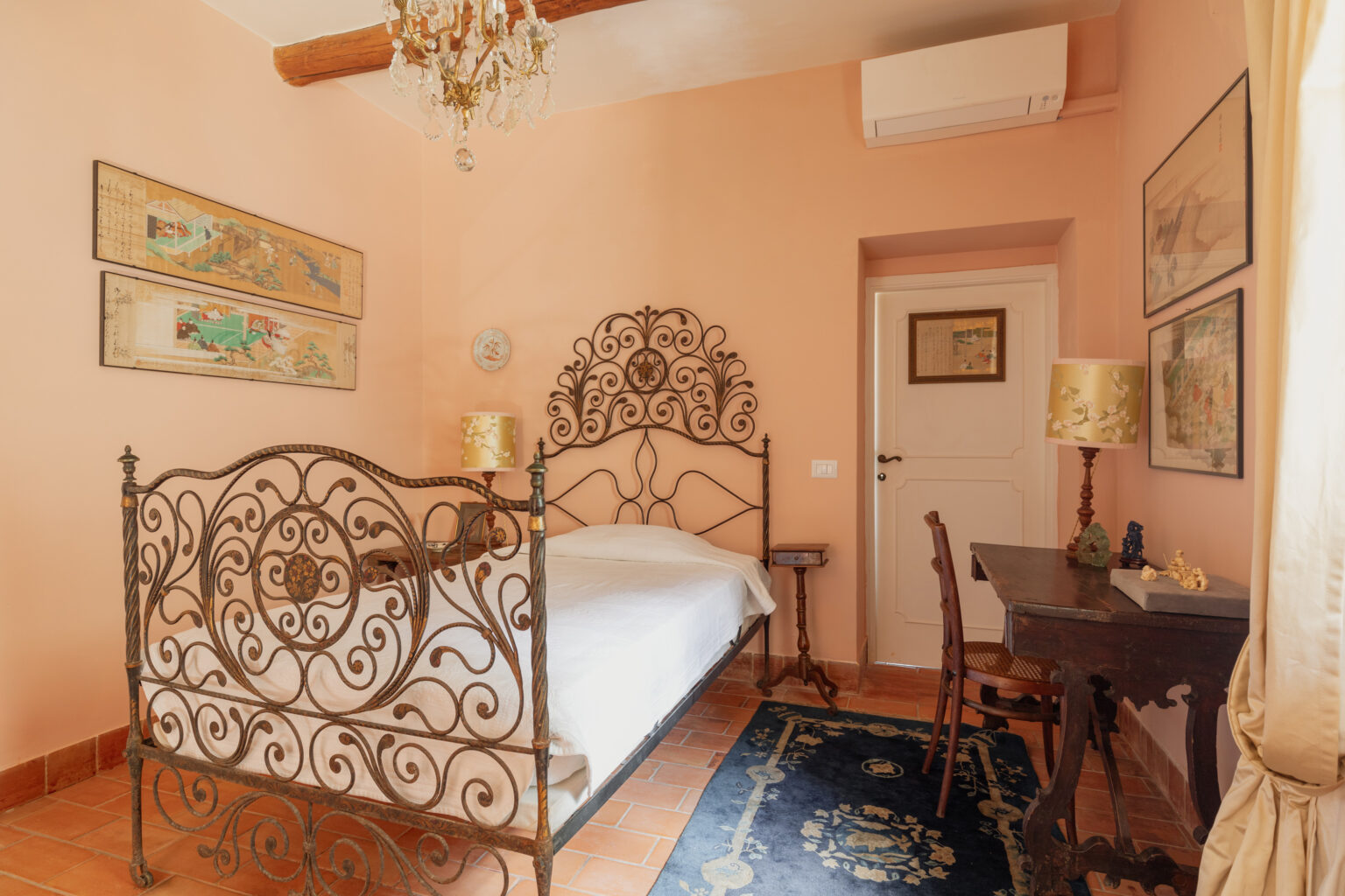 Cozy bedroom featuring an ornate wrought iron bed, peach-colored walls, and a vintage wooden desk. The room is adorned with framed artwork, an elegant chandelier, and a decorative lamp, creating a warm and inviting atmosphere. | Only on SopranoVillas
