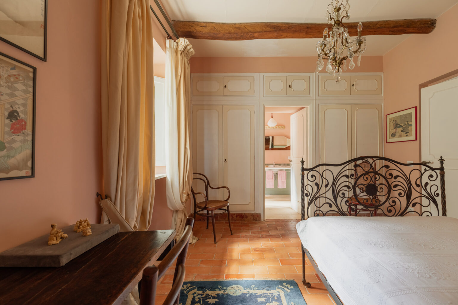 Cozy and charming bedroom featuring a wrought iron bed, wooden beams, and terracotta flooring. The room is decorated with light pink walls, elegant curtains, and framed artwork, creating a warm atmosphere. A wooden table and a chair add functionality, while a doorway leads to a stylish kitchen area in the background. | Only on SopranoVillas