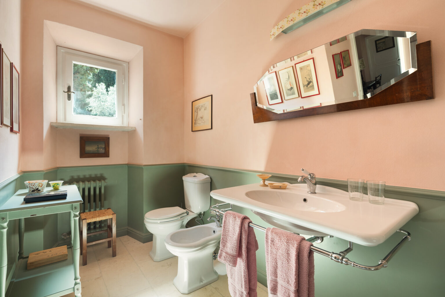 Bright and airy bathroom featuring pastel pink walls and green wainscoting, equipped with a modern sink, toilet, and bidet. The space includes a large mirror, decorative artwork, and a window providing natural light, enhancing the inviting atmosphere. | Only on SopranoVillas