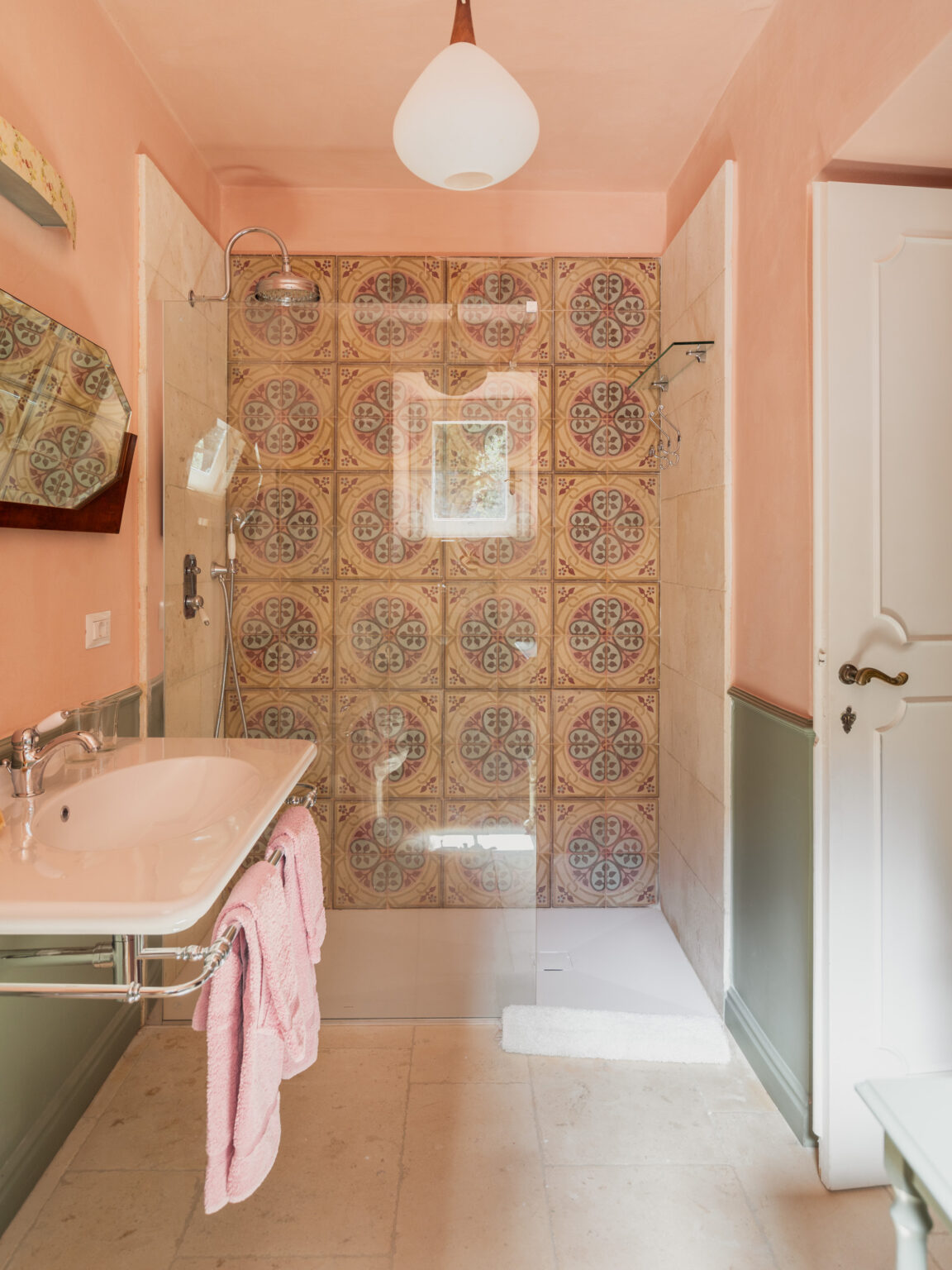 Stylish bathroom featuring a glass shower, decorative tile wall, modern sink with chrome fixtures, and soft pink towels hanging on a rack, complemented by a warm peach wall color and elegant lighting. | Only on SopranoVillas