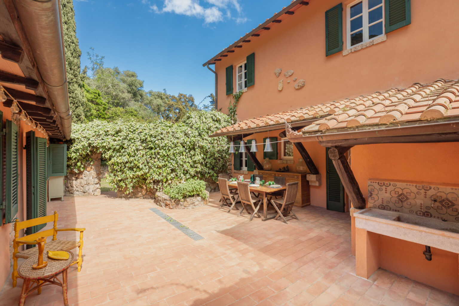 Charming outdoor patio of a rustic house featuring terracotta tiles, a wooden dining set, and lush greenery. The space is adorned with hanging lights and a stone sink, perfect for al fresco dining in a serene setting. | Only on SopranoVillas