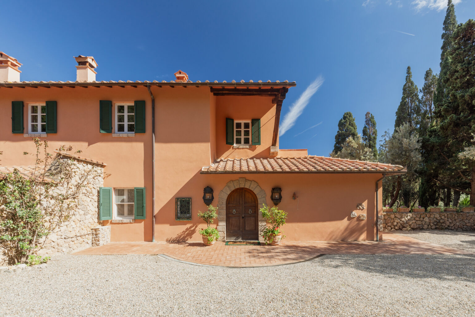 Charming Mediterranean-style house with a terracotta roof, green shutters, and a wooden front door, set against a clear blue sky and surrounded by lush greenery. The gravel driveway leads to the entrance, showcasing the rustic stonework and landscaped details. Ideal for showcasing architectural beauty and outdoor living. | Only on SopranoVillas