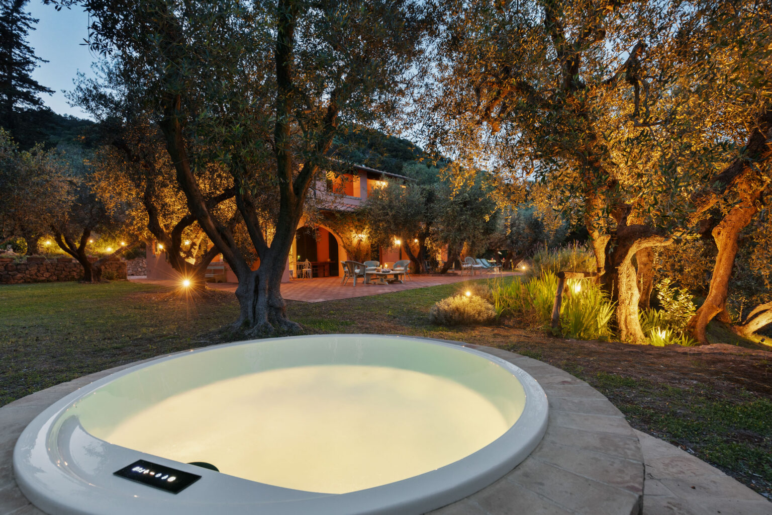 Luxurious outdoor hot tub surrounded by illuminated olive trees and a beautifully landscaped garden, with a glimpse of a charming villa in the background during twilight. | Only on SopranoVillas