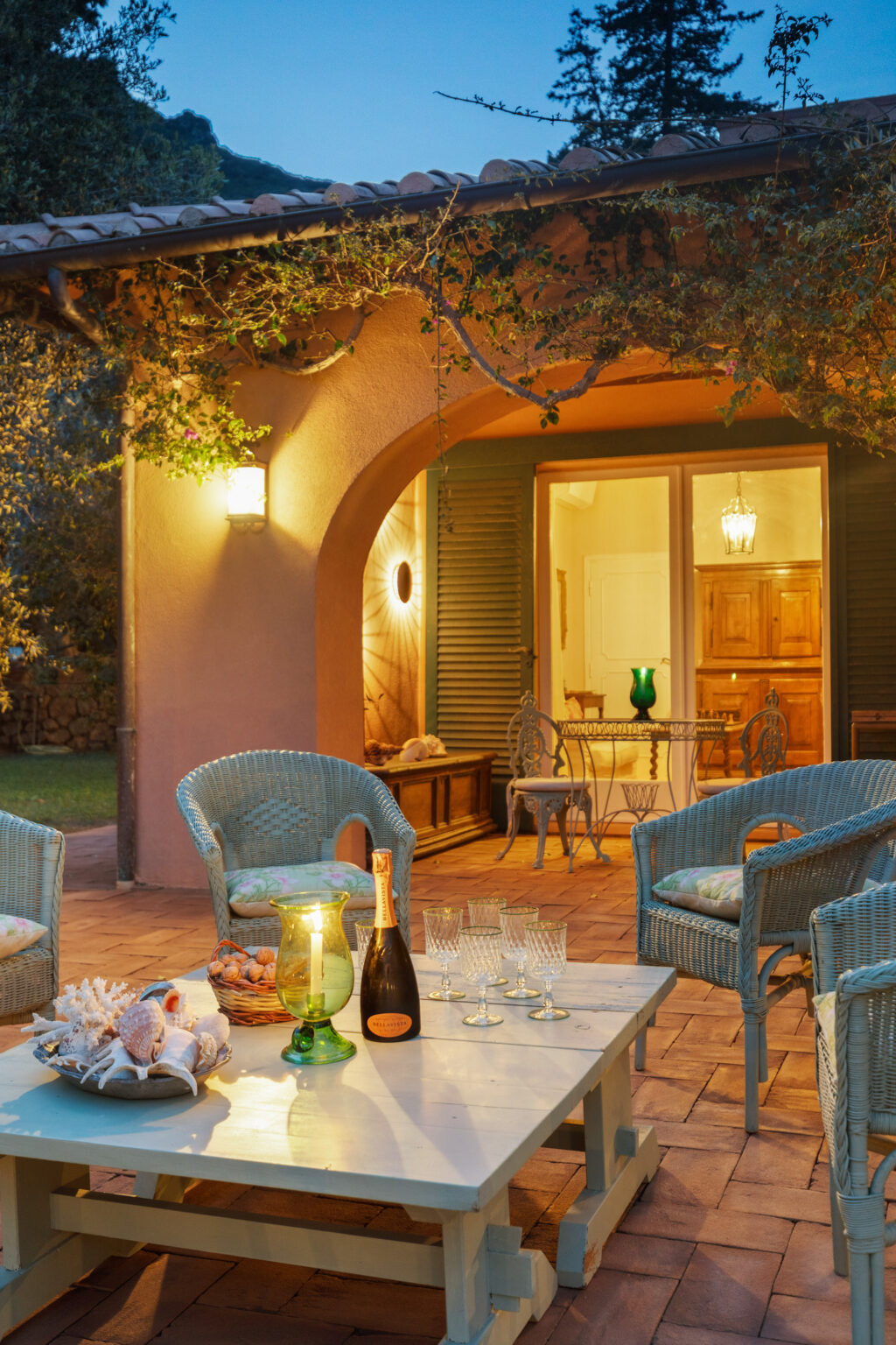Cozy outdoor patio setting at dusk featuring blue wicker chairs, a white coffee table adorned with seashells, a green glass lantern, a bottle of champagne, and crystal glasses, with a beautifully lit doorway leading into a warm interior space. | Only on SopranoVillas